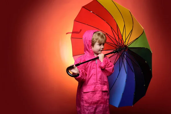 Niño pequeño con paraguas de color arco iris aislado sobre fondo rojo. Los niños anuncian sus productos y servicios. Lindo niño pequeño usando ropa de otoño en el día de lluvia de otoño. Niño bajo la lluvia . — Foto de Stock