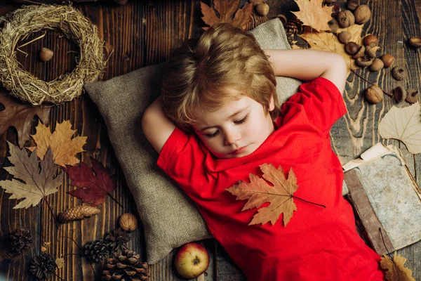 Söt liten barn pojke är redo för hösten. Barn annonsera dina produkter och tjänster. Blond liten pojke vilar med blad på mage ligger på trägolv i höstlöv. — Stockfoto