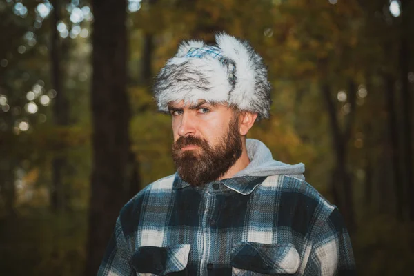 A young guy in the hat, traveller, hipster standing in the woods. Hiking, Forest, Journey. Portrait of a young bearded man in warm ethnic hat on autumn forest background. Autumn concept. — Stock Photo, Image