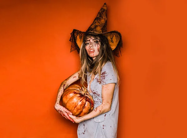Model s krvavou rukou a krev na tváři. Šťastná mladá žena halloween kostým s kloboukem stojící nad červeným pozadím. Čarodějnice Halloween s dýní. Šaty na Maškarní Halloween. — Stock fotografie