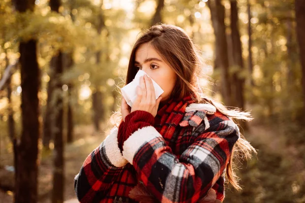 Young woman with nose wiper near autumn tree. Sick girl with runny nose and fever. Showing sick woman sneezing at autumn park. Young woman having flu and blowing her nose. — Stock Photo, Image