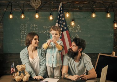 Ebeveynler sınıf ABD bayrağı ile kara tahta arka plan ile evlat. Amerikan ailesinin oğlu ve ABD bayrağı ile masa başında oturmak. Vatansever eğitim kavramı. Anne oğlu Amerikan gelenekleri oynarken öğretim.