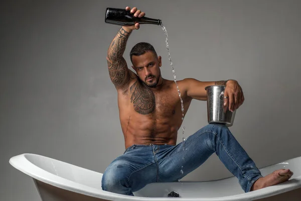 Weinfestkonzept. Sekt- oder Weinfest. Schöner brutaler Mann gießt Champagner in die Badewanne auf grauem Hintergrund. — Stockfoto