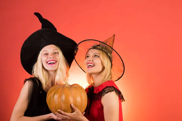 Deux jeunes femmes heureuses en robes noires et rouges, costumes sorcières Halloween sur fond orange. Deux filles sexy Halloween avec citrouille. Design festif d'Halloween. Des gens heureux. S'amuser . — Photo