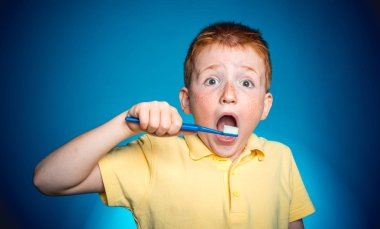 Çocuk temiz dişler diş fırçası ile. Üzerinde mavi arka plan izole diş fırçası ile evlat. Komik surat ile Kızıl saçlı çocuk fırçalarken diş fırçası ile. Mutlu çocuk çocuk çocuk diş fırçalama.