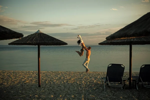 El hombre lleva a la mujer, pareja feliz de vacaciones. Pareja en el amor bailando, divirtiéndose, mar y horizonte fondo. Pareja enamorada parada en la playa, a orillas del mar. Luna de miel, concepto de recién casados . — Foto de Stock