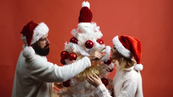 Pareja cariñosa en los sombreros de Año Nuevo adorna un árbol de Navidad sobre un fondo rojo. Hombre barbudo y chica rubia se están preparando para celebrar el año nuevo. Concepto de celebración del nuevo año . — Vídeos de Stock