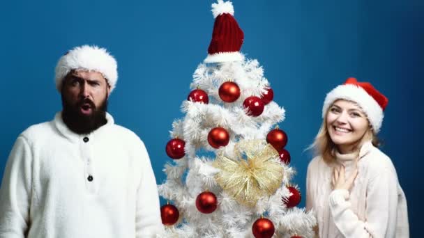 新しい年の帽子のひげを生やした男と赤い帽子の女は クリスマス ツリーの近くに立つし 感情を示します 新年のお祝いのコンセプト — ストック動画