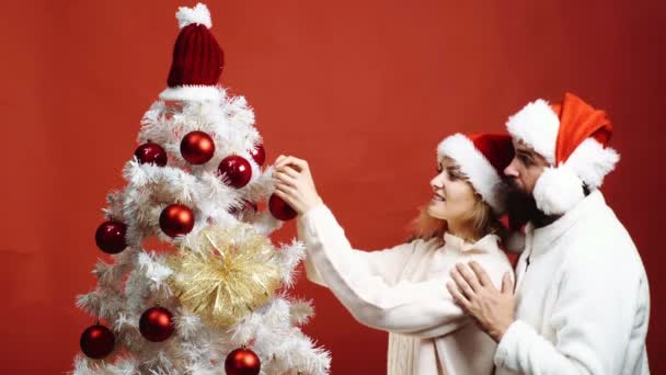 Skäggig man bär en röd hatt omfamnar en kvinna som pryder en julgran. Underbara par dekorerar en julgran. Begreppet firandet av nyår. — Stockvideo