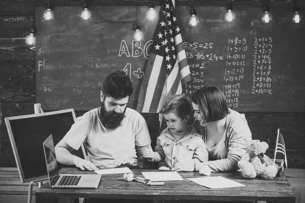 Niño con padres en el aula con bandera de EE.UU., pizarra en el fondo. La familia americana se sienta en el escritorio con su hijo y su bandera. Padres enseñando a tocar a sus hijos tradiciones americanas. Concepto de educación patriótica . — Foto de Stock