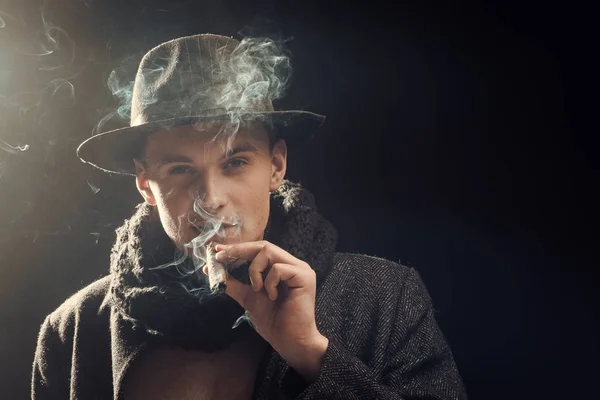 Hombre con abrigo, sombrero fumando cigarro, fondo oscuro. Macho en cara misteriosa, detective, investigador, agente. El tipo de traje anticuado parece misterioso con cigarro y humo. Concepto de detective Vintage . —  Fotos de Stock