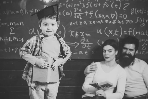 Parents listening their son, checking mistakes with book chalkboard on background. Boy presenting his knowledge to mom and dad. Smart child in graduate cap like to perform. Preparing for exam concept.