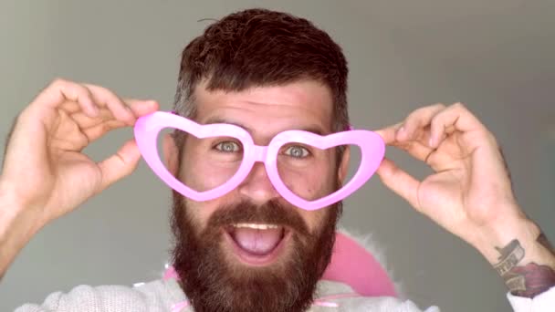 Verrückter Mann mit lustiger hipster pinkfarbener Sonnenbrille und komischem rosa Hut. Lustiger Hipster, der vor grauem Hintergrund in die Kamera blickt. Ausdruck und Personenkonzept - Mann mit lustigem Gesicht vor grauem Hintergrund. — Stockvideo