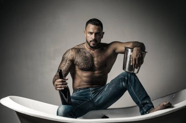 Şampanya kutlaması. Gri arka planda dövmeli yakışıklı bir adam. Seksi gece partisi, genç aşıklar. Kot pantolonlu moda adamı bacakları.
