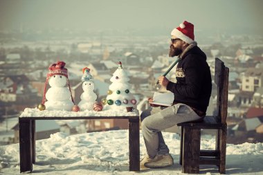 Hippi kalem ve defter sandalyede oturuyor. Noel Baba adam kış gününde yazma. Noel ve yeni yıl tatilleri kutlama. Kardan adam ve kar xmas ağacı ahşap masanın üzerinde. Öğrenme ve eğlenceli kavramı.
