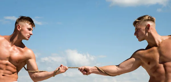 Lleno de fuerza. Gemelos competidores con cuerpos musculares. Gemelos atléticos en lados opuestos. Los hombres fuertes tiran de la cuerda con fuerza muscular de la mano. Los hombres muestran su fuerza contra los competidores —  Fotos de Stock