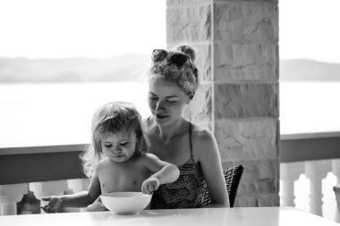 Çocuk kendini doğal zemin üzerine sütunlarla açık terasta annesiyle masada oturan yiyor.