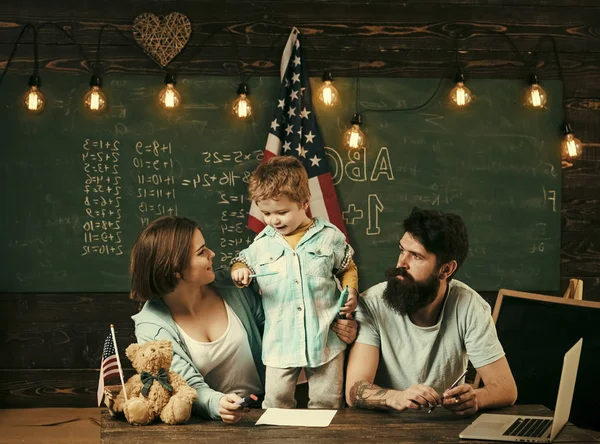 Amerykańska rodzina siedzieć przy biurku z flagą syn i usa. Dziecko z rodzicami w klasie z flagą usa, tablica na tle. Koncepcja edukacji patriotycznej. Rodziców, uczy syna amerykańskiej tradycji gry. — Zdjęcie stockowe