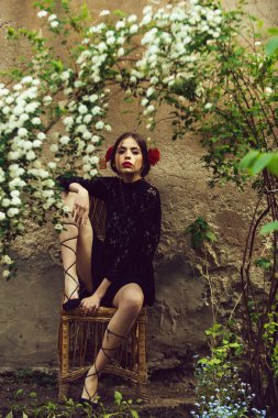 Sevimli kadın ya da güzel bir kız, genç moda model kırmızı dudaklar, saç ve bush ile beyaz, altında hasır sandalye üzerinde oturan siyah dantel elbise içinde gül ile bej açık duvar çiçek çiçek açması. Bahar
