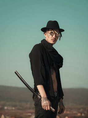 Savaşçı güneş gözlüğü, siyah şapka, açık giysili dövmeli gövde. Samuray ve Japonya silah. Dövüş sanatları kavramı. Konsantrasyon ve zen. Mavi gökyüzü üzerinde katana kılıcı duran adamla.