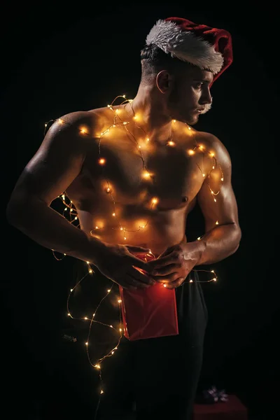 大人のための贈り物で新年パーティー ストリップでガーランドに筋肉のついた体 クリスマス サンタ衣装で若い男とサンタ — ストック写真