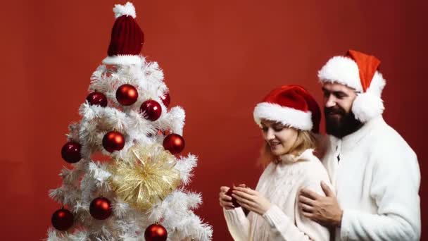 Pareja cariñosa con caras felices cumplir el Año Nuevo cerca del árbol de Navidad. Pareja joven en los sombreros de Año Nuevo adornan el árbol de Navidad sobre el fondo rojo. Esposo abraza a una mujer que viste un árbol de Navidad . — Vídeo de stock