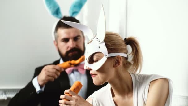 穿着西装的男人和一个穿着兔子皮面膜的女人吃胡萝卜在白色的背景。花花公子概念. — 图库视频影像