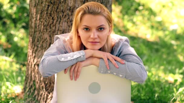 Девушка сидит в парке, опираясь на ноутбук. Девушка отдыхает в парке с ноутбуком . — стоковое видео