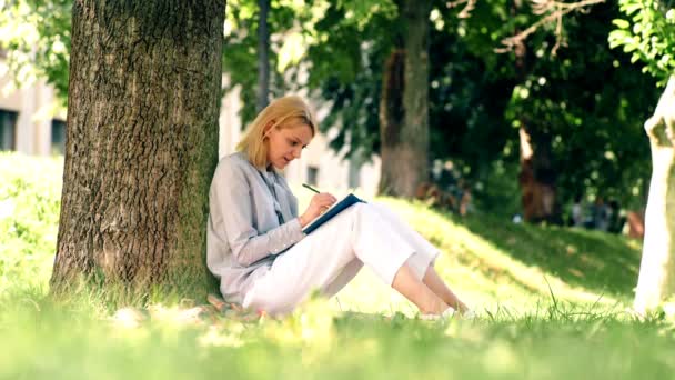 Chica hace notas en un cuaderno mientras está sentado en el parque en la hierba. Estudiante está estudiando en un parque. — Vídeos de Stock