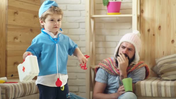 Boy and father play physician and patient. The boy in the doctors suit treats a bearded man in a knitted hat. Concept of a medical case. — Stock Video