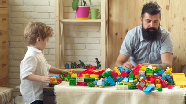 Padre e hijo con caras felices crean construcciones coloridas con ladrillos de juguete. El niño y su padre juegan con el diseñador lego. Concepto de familia e infancia. — Vídeo de stock