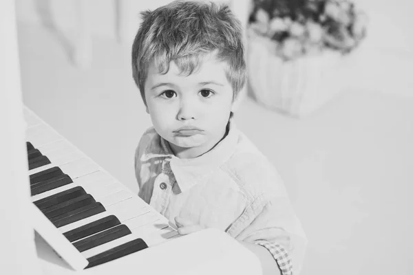 Niño sentado cerca del teclado del piano, fondo blanco. Niño lindo y adorable pone el dedo en el teclado del piano. Los niños pasan el ocio cerca del instrumento musical. Elite concepto de infancia . — Foto de Stock
