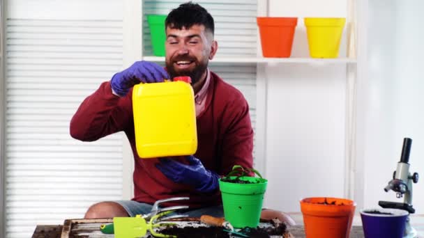 L'uomo barbuto sorridente tiene in mano un barattolo di plastica e in un'altra mano tiene un vaso con una pianta. Il concetto di giardinaggio . — Video Stock