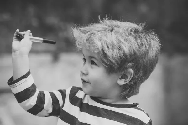 Child or boy with smiling face wears striped clothes. Childhood concept. Cute, adorable kid or son with untidy blonde hair on light background, defocused. Boy holds or plays with pen or marker in hand