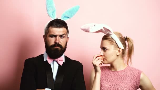 Bearded man with ears hare and blond woman with ears hare eating an apple on a pink background. A woman with rabbit ears offers an apple to her husband with rabbit ears.