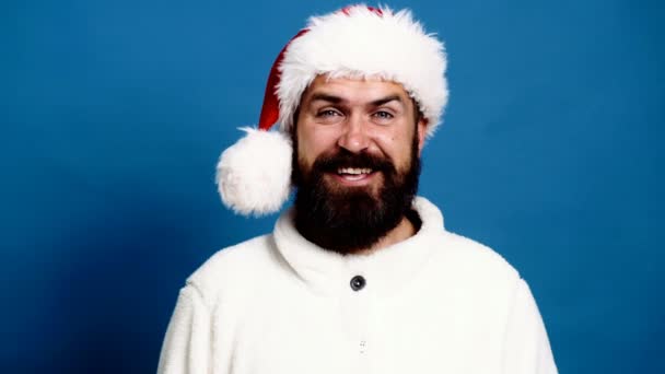 Bearded man wearing a red New Year hat moves his eyebrows and smiles on a blue background. Funny Santa Claus. Concept of new year mood. — Stock Video