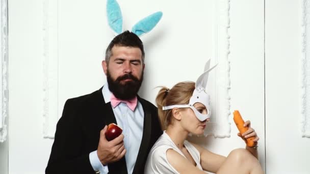 Un homme barbu portant un costume et des oreilles de lapin mange une pomme avec une femme vêtue d'un masque en cuir qui mange une carotte. Couple à la mode mange des fruits sur fond blanc . — Video