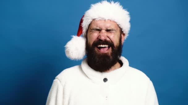 Ein bärtiger Mann mit Neujahrsmütze lächelt aufrichtig auf blauem Grund. Lustiger Weihnachtsmann auf blauem Hintergrund. Konzept der Weihnachtsstimmung. — Stockvideo