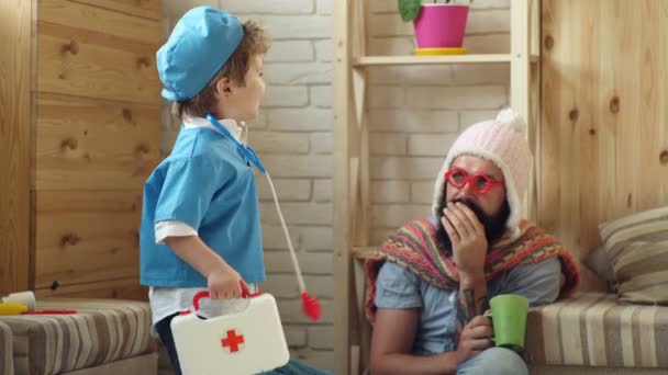 Father and son playing doctor, healthcare and treatment concept. Emergency medical specialist visiting patient at home. Man with long beard in knitted white hat coughing due to flu. — Stock Video