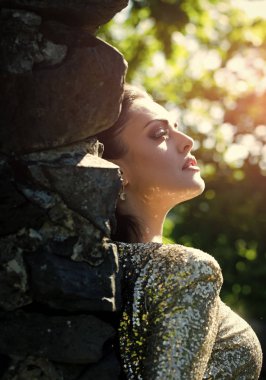 Kadın taş duvara güneşli günde poz. Kız doğal arka plan bulanık. Altın pul ile parti elbise moda modeli. Tatilleri, doğum günü, yıldönümü kutlamaları. Güzellik ve doğa