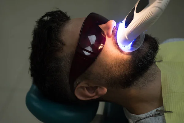 Examen du jeune homme par un dentiste sur fond flou. Bel homme européen sourire avec des dents saines blanchissant. Dentiste féminine vérifiant les dents du patient avec miroir dans la clinique dentaire moderne. — Photo