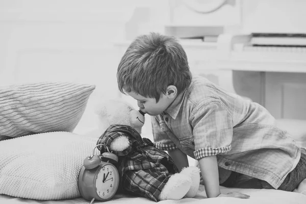 Child in bedroom kiss toy in nose. Good night concept. Boy with happy face puts favourite toy on bed, wishing sweet dreams. Kid put plush bear near pillows and alarm clock, luxury interior background. — Stock Photo, Image