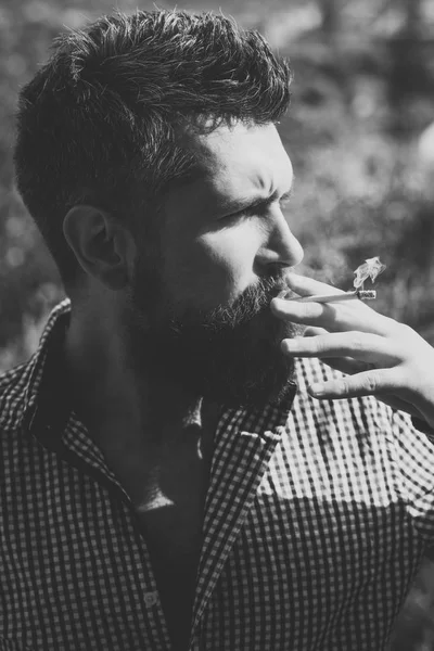 Joven barbudo hipster con barba y bigote en la cara seria fumar cigarrillos al aire libre. hombre fumando cigarrillo. estilo de vida poco saludable y mal hábito de chico con adicción. peluquería y peluquería — Foto de Stock