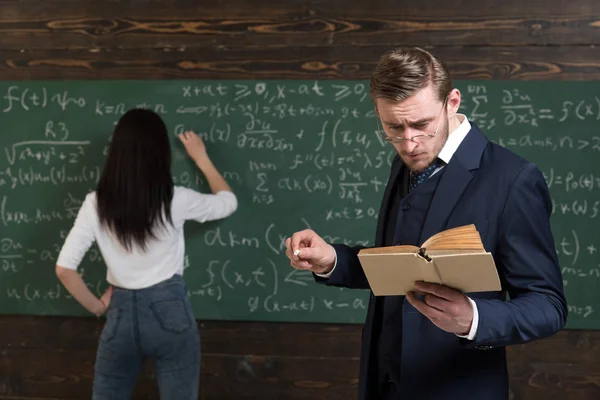 上学日。戴眼镜的老师从课本读到女生的问题陈述。学习数学使我能思考清楚。后视女子在黑板上写方程。做求和 — 图库照片