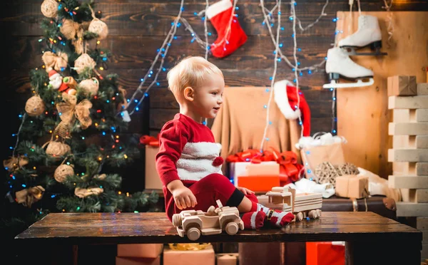 Счастливый малыш, одетый в зимнюю одежду, думает о Санте возле рождественской елки. Рождество для детей. Зимний вечер дома. Улыбающийся ребенок подглядывает из-за елки в гостиной . — стоковое фото