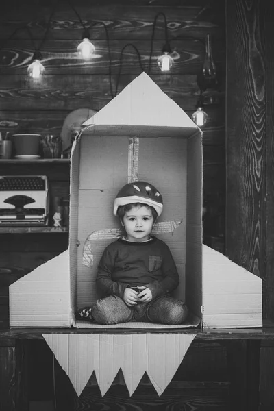 Kid in helmet sit in cardboard hand made rocket. Child boy play cosmonaut, astronaut. Childhood concept. Boy play at home with rocket, little cosmonaut sit in rocket made out of cardboard box.