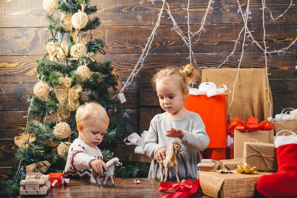 Niedliches kleines Mädchen schmückt den Weihnachtsbaum drinnen. Frohe Weihnachten. Porträt Kind mit Geschenk auf Holzgrund. Lächelndes Kind lugt hinter Weihnachtsbaum im Wohnzimmer hervor. — Stockfoto