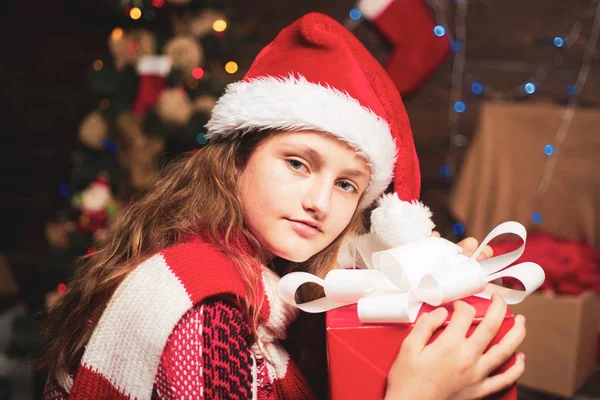 Vánoční dárek teenagera. Vánoční děti. Malé radostné holčičky na sobě zimní oblečení přemýšlet o Santa u vánočního stromu. Šťastný, malé teenager v Santa čepice s současnosti mají Vánoce. — Stock fotografie