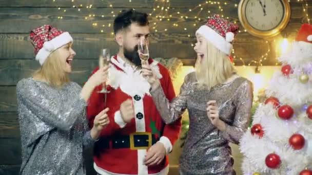 Бородатий чоловік в костюмах Сантас і дві дівчини в новорічних капелюхах п'ють шампанське і спалюють блискітки на тлі новорічних пейзажів. Концепція новорічного настрою . — стокове відео