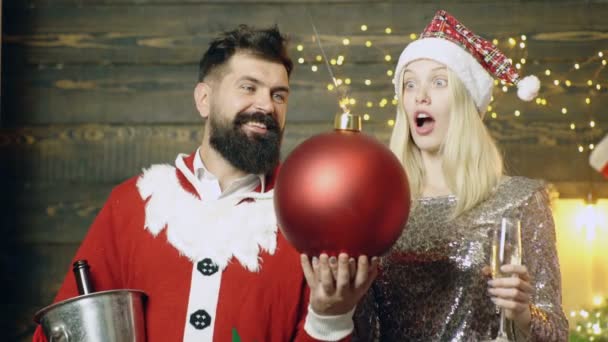 Weihnachtsbombe. Kreativboom. Bärtiger Mann im Weihnachtsmannkostüm und blondes Mädchen mit Neujahrsmütze blasen eine Bombe, die in ihren Händen hält. Mann mit einem Mädchen bläst eine Silvesterbombe. — Stockvideo